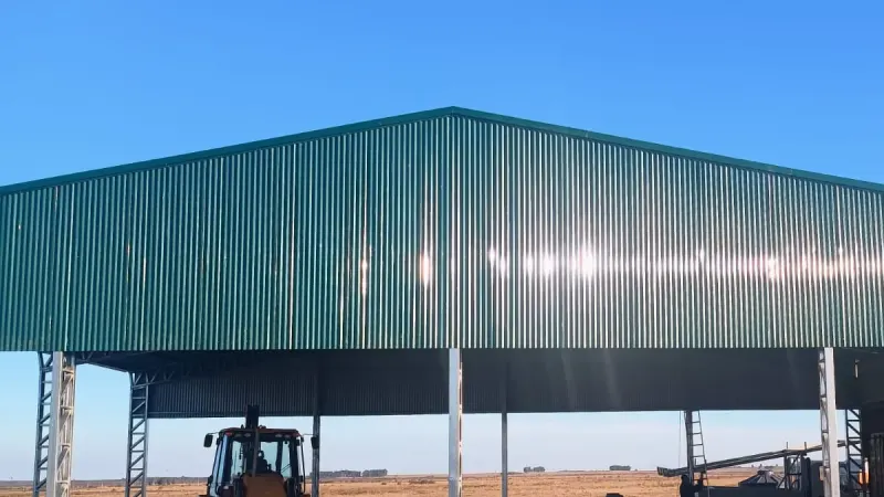 Vista parcial: tinglado abierto de chapa verde con una retroexcavadora debajo en campo abierto