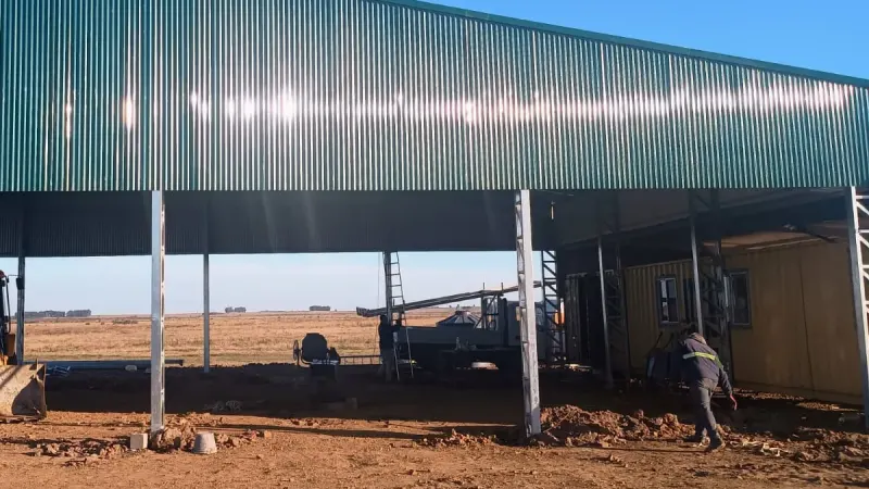 Otro ángulo: tinglado abierto de chapa verde con una retroexcavadora debajo en campo abierto